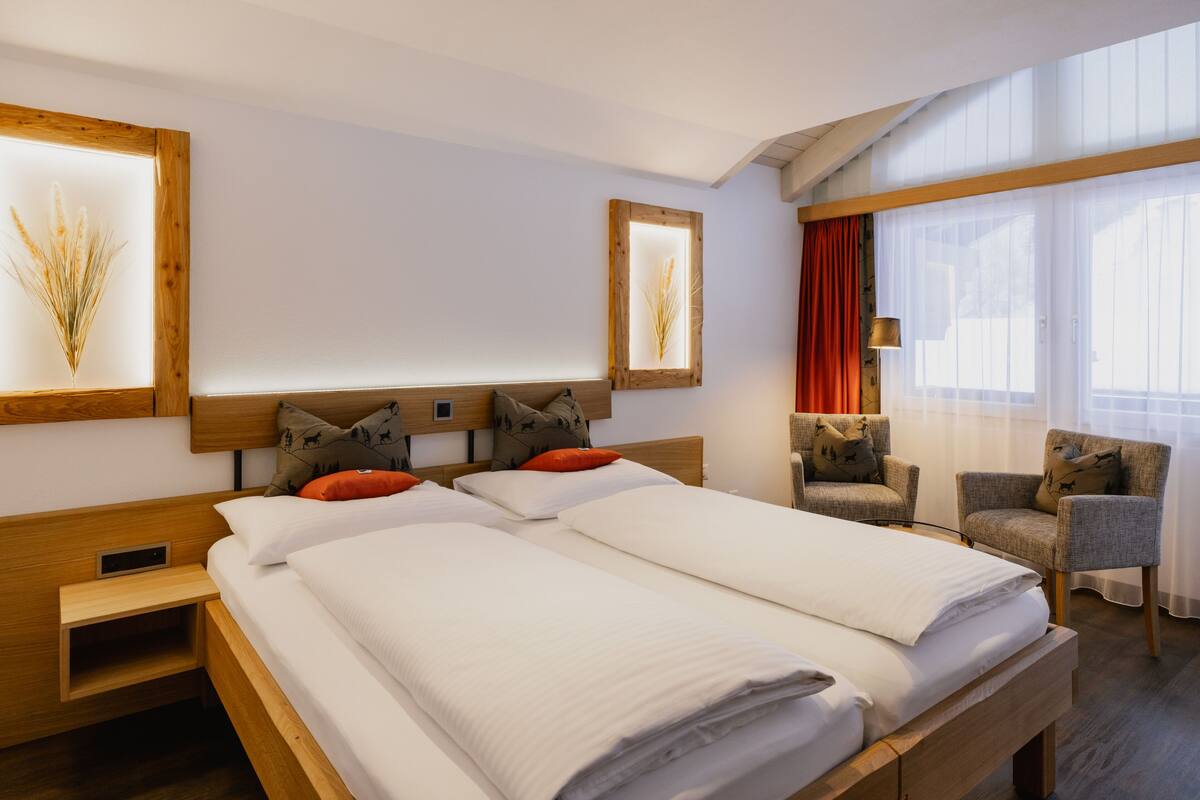 Een hotelkamer met een houten bed, twee nachtkastjes, een stoel en een groot raam met gordijnen.