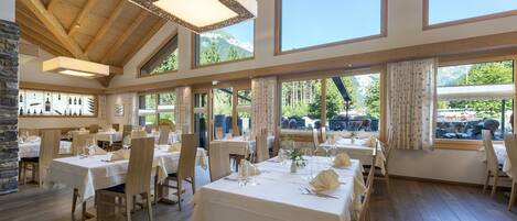 A restaurant with multiple tables set for dining, large windows offering views of the outdoors, and a wooden ceiling structure.