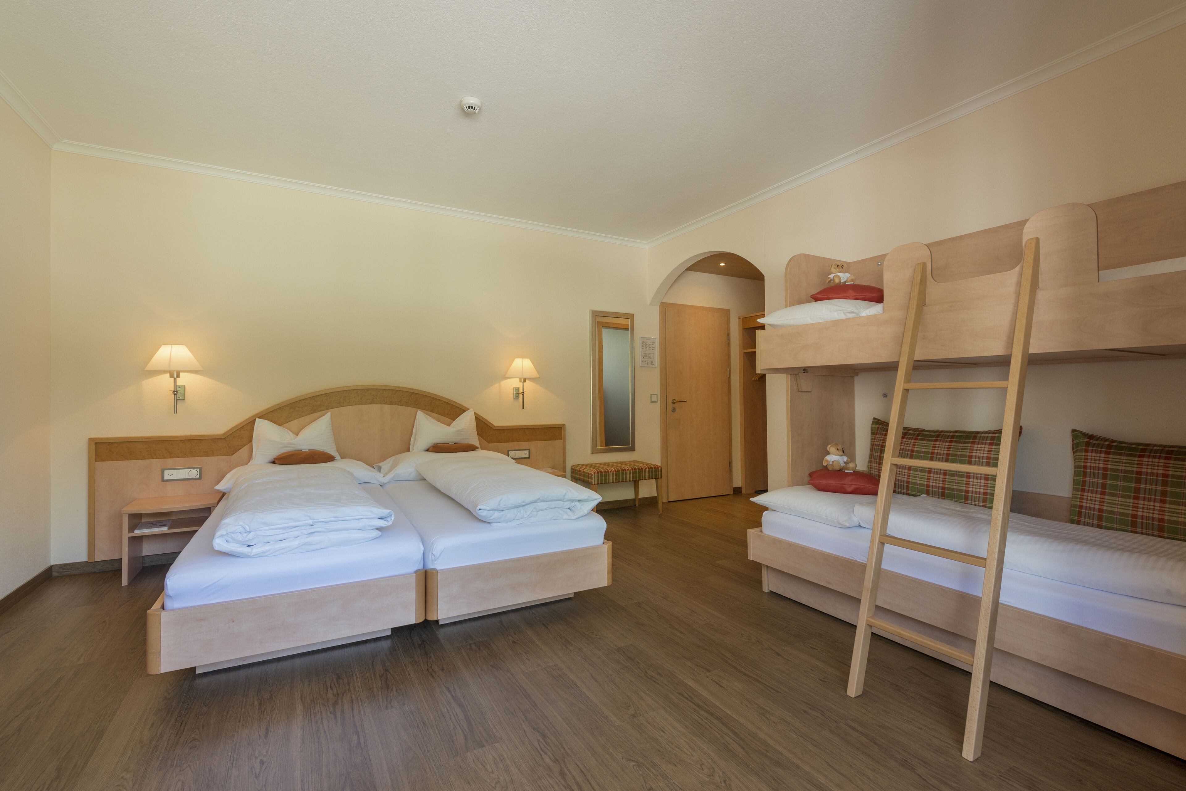 A hotel room with two beds, a bunk bed, wooden flooring, and a small bedside table with a lamp.