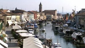 Un canale fiancheggiato da barche ed edifici, con una torre visibile in lontananza e una passeggiata pedonale.
