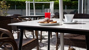 Zona pranzo all'aperto con sedie in vimini e un tavolo apparecchiato con un piatto di cibo, un bicchiere di bevanda rossa e una tazza bianca.