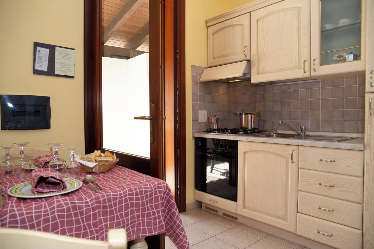 A kitchen with wooden cabinets, a stove, and a sink. There is a dining table with a tablecloth and cutlery.
