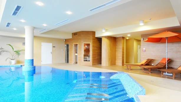 En moderne indendørs swimmingpool med et klart blåt vandelement, et tilstødende loungeområde med træliggepladser og en orange parasol, samt en trævæg med en glasdør, der fører til et spa-område.