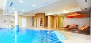 Una piscina interior moderna con una fuente de agua azul cristalina, zona de estar contigua con tumbonas de madera y un paraguas naranja, y una pared de madera con una puerta de cristal que da a un área de spa.