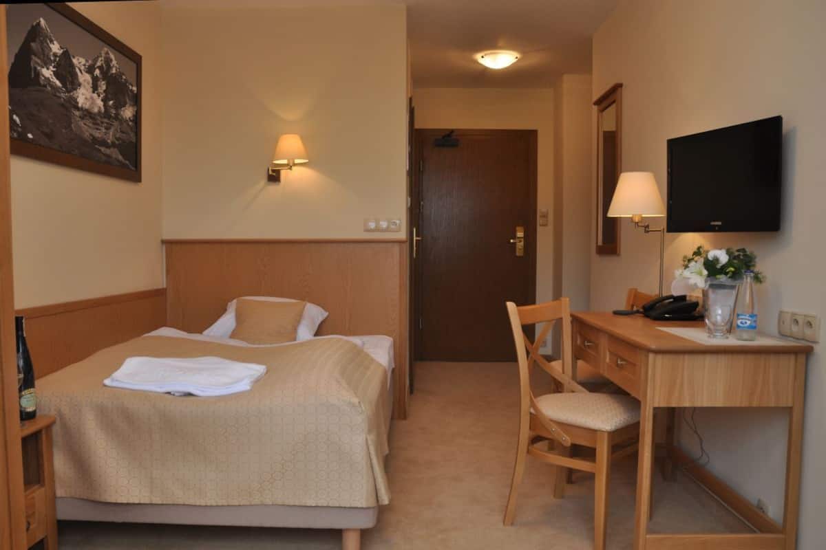 A hotel room with a bed, a desk, a chair, a television, and a wall-mounted lamp.