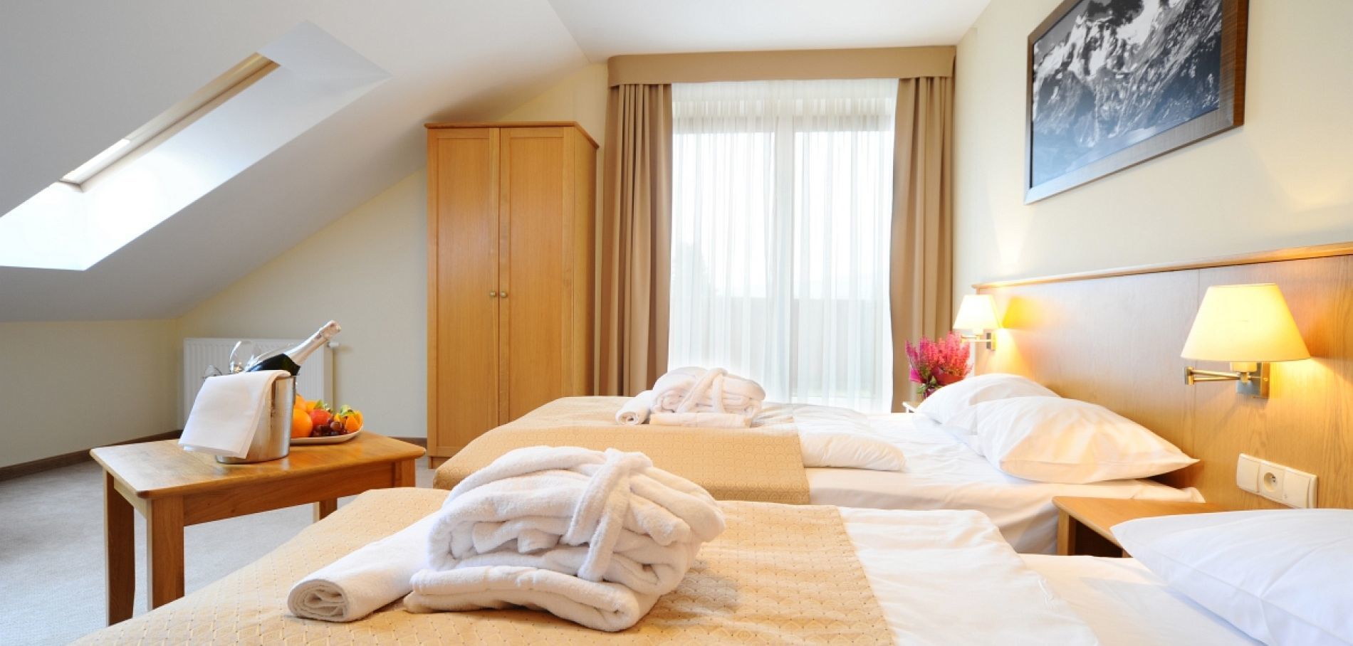 A hotel room with two beds, a wooden wardrobe, a desk with a bottle and fruit, and a painting on the wall.