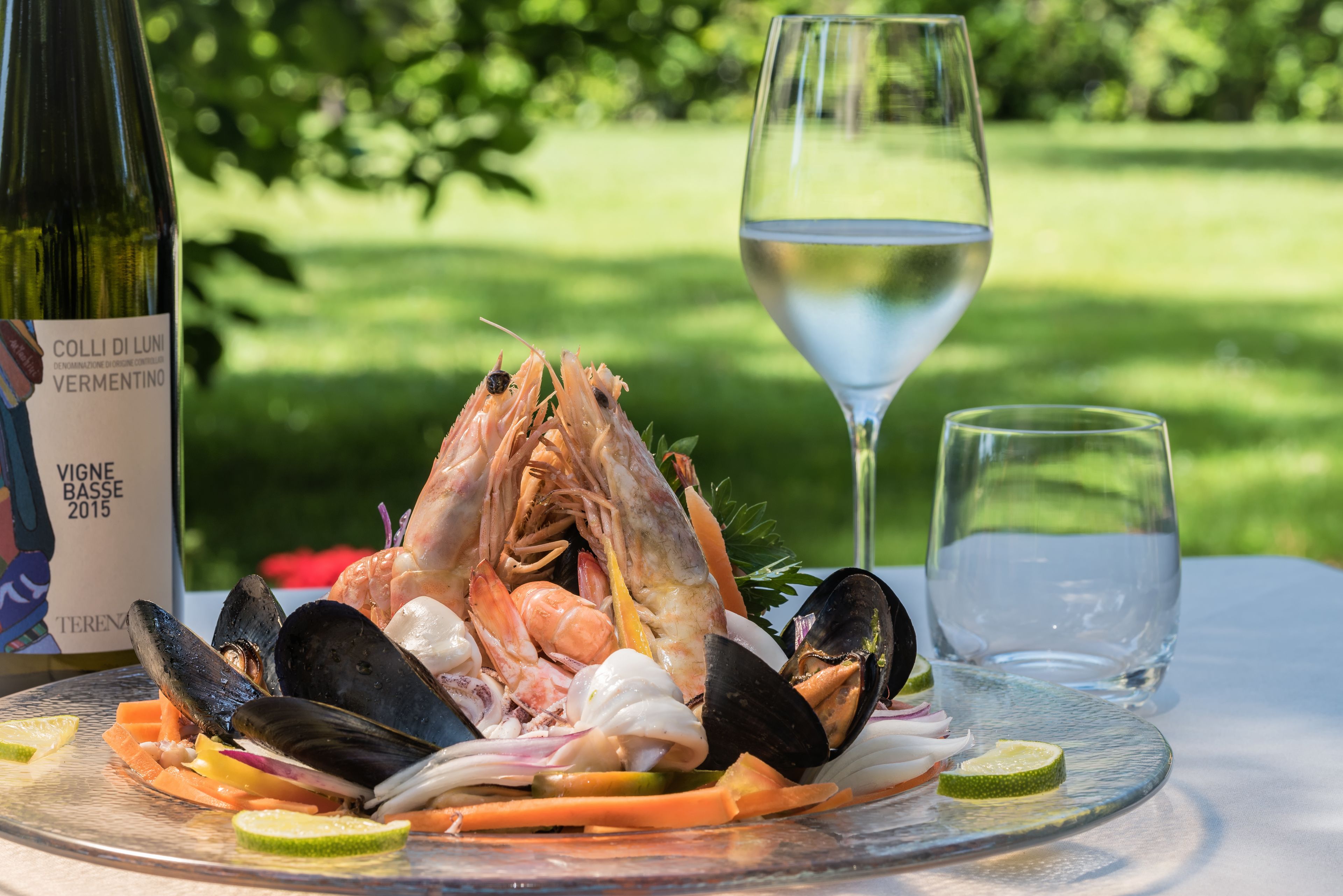 A seafood platter with mussels, shrimp, and other shellfish, accompanied by a bottle of wine and a glass of water, set on a table outdoors.