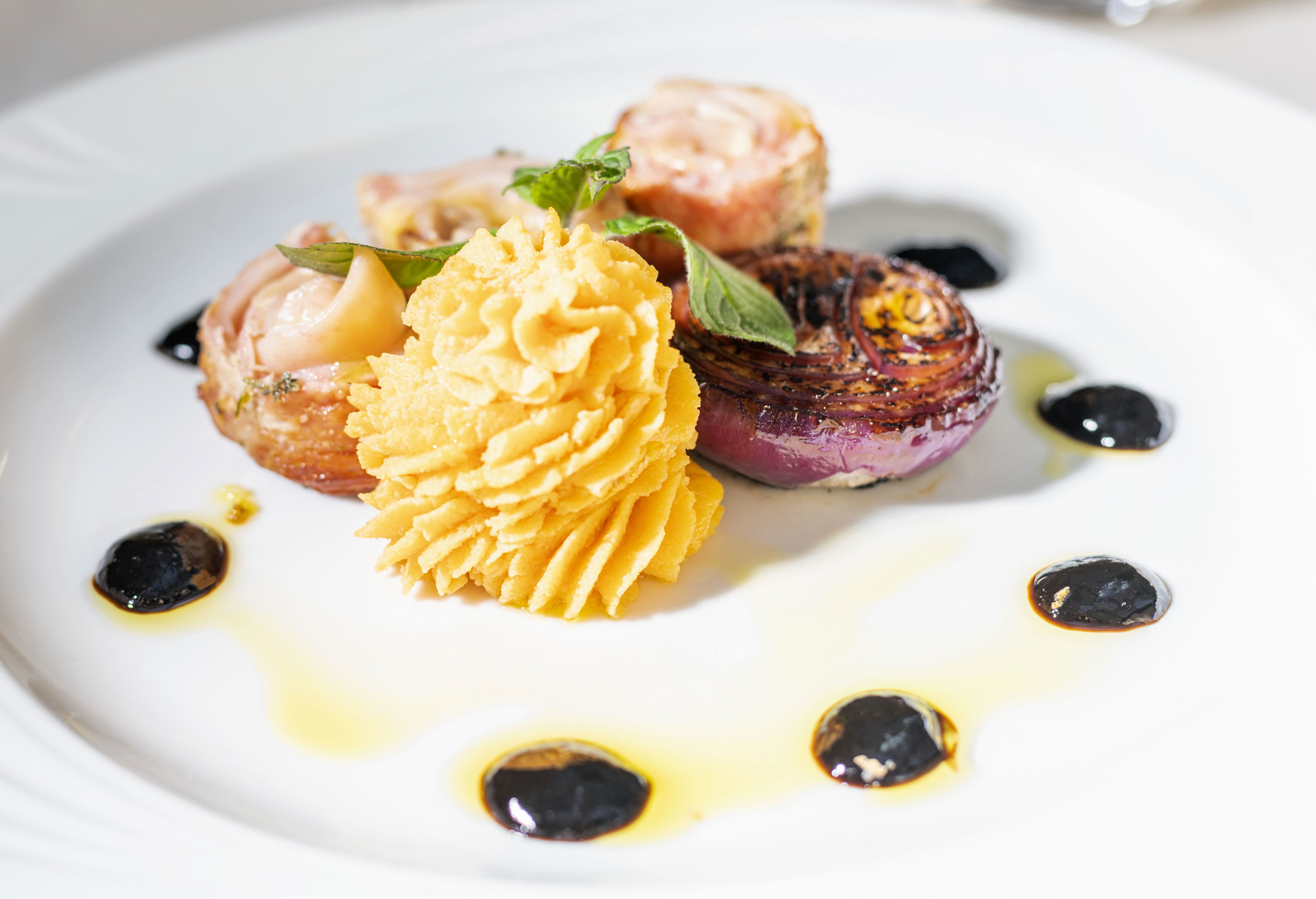 Un plato blanco con una composición de alimentos que incluye un postre amarillo, un plato a la parrilla y un pequeño pastel, con salpicaduras de salsa negra y amarilla.