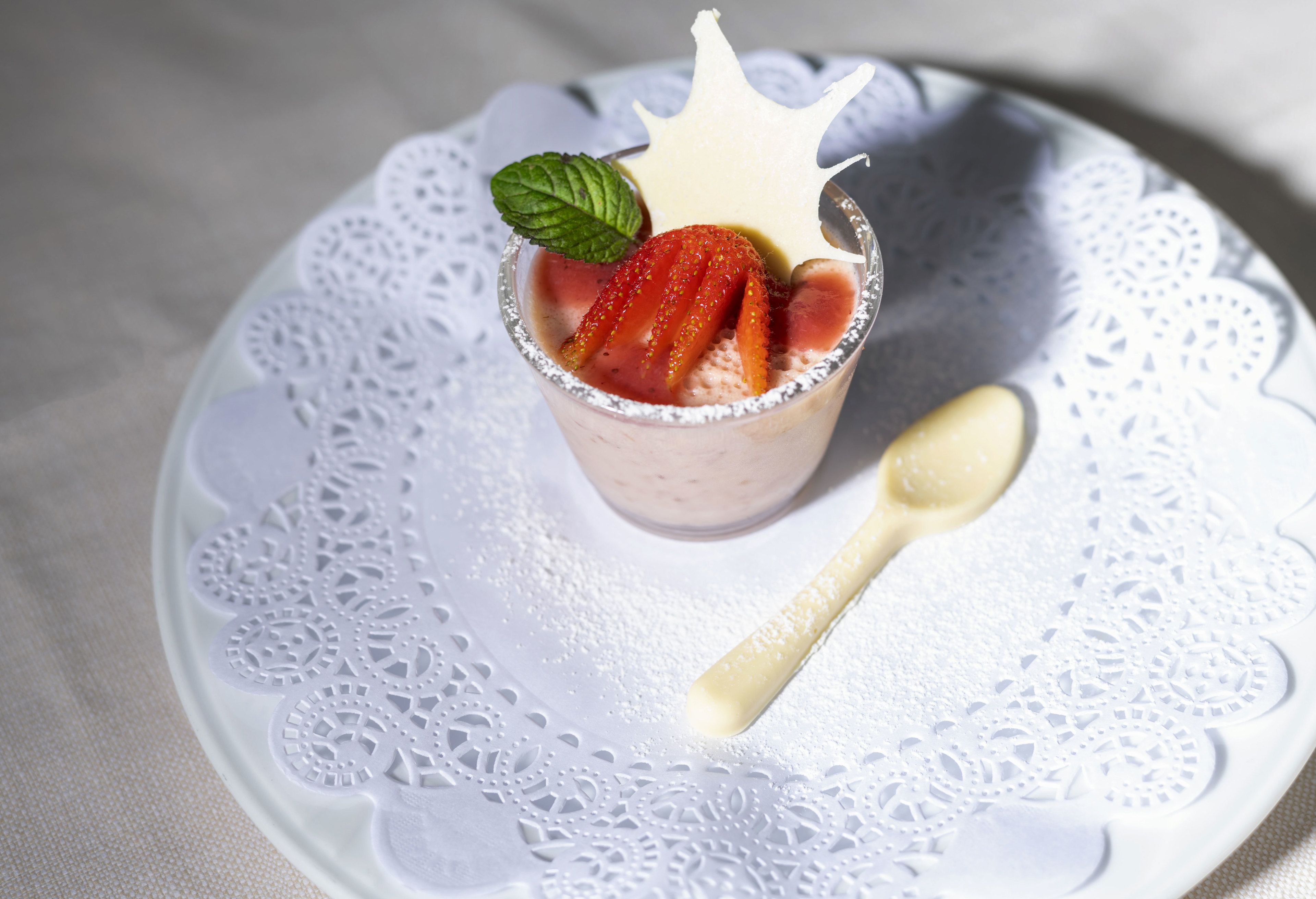 A dessert in a clear glass with strawberries and a mint leaf on top, served on a white plate with a lace doily underneath.