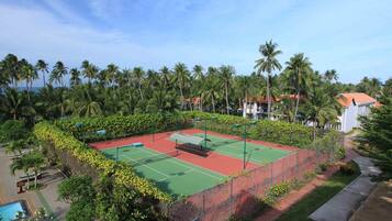 A tennis court surrounded by lush greenery and palm trees, with a pool and a building in the background.