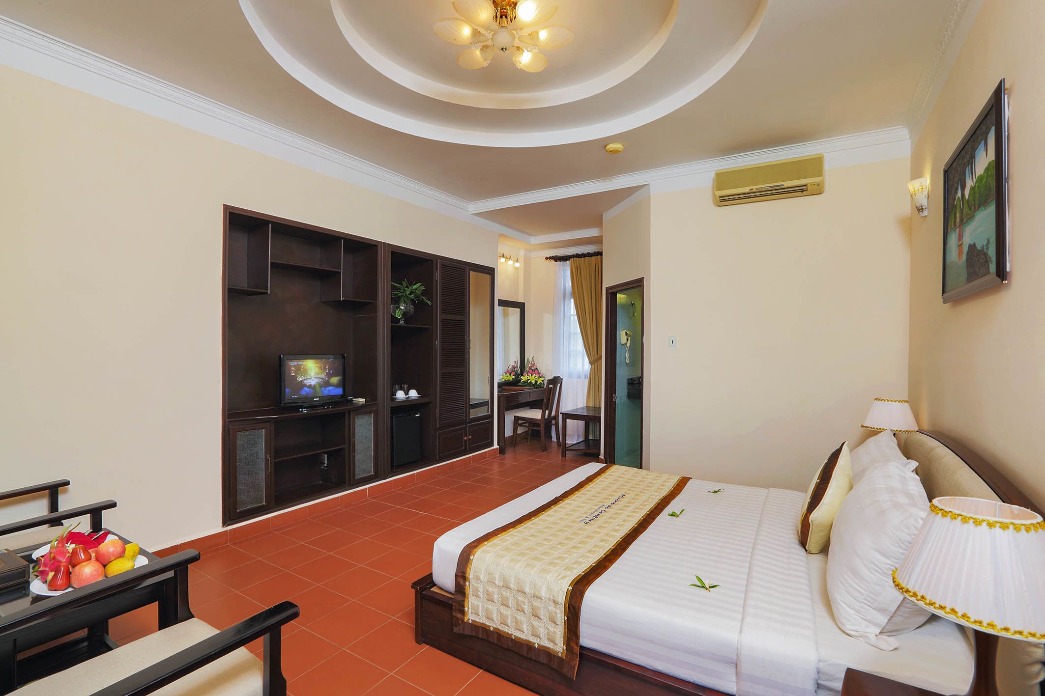 A hotel room with a bed, a desk with a television, a small table with fruit, and a wall-mounted air conditioner.