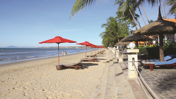 Ein Strand mit Liegestühlen, roten Sonnenschirmen, Palmen und freiem Blick auf den Ozean.
