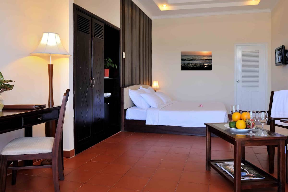 Ein Hotelzimmer mit Bett, Schreibtisch mit Lampe, Stuhl, Esstisch mit Obst und Gläsern sowie Fernseher an der Wand.