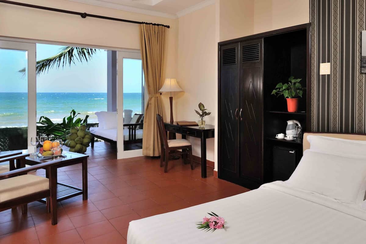 Ein Hotelzimmer mit Bett, Schreibtisch, Stuhl, einem kleinen Tisch mit Obst und Gläsern, einer Topfpflanze und Blick auf den Ozean.