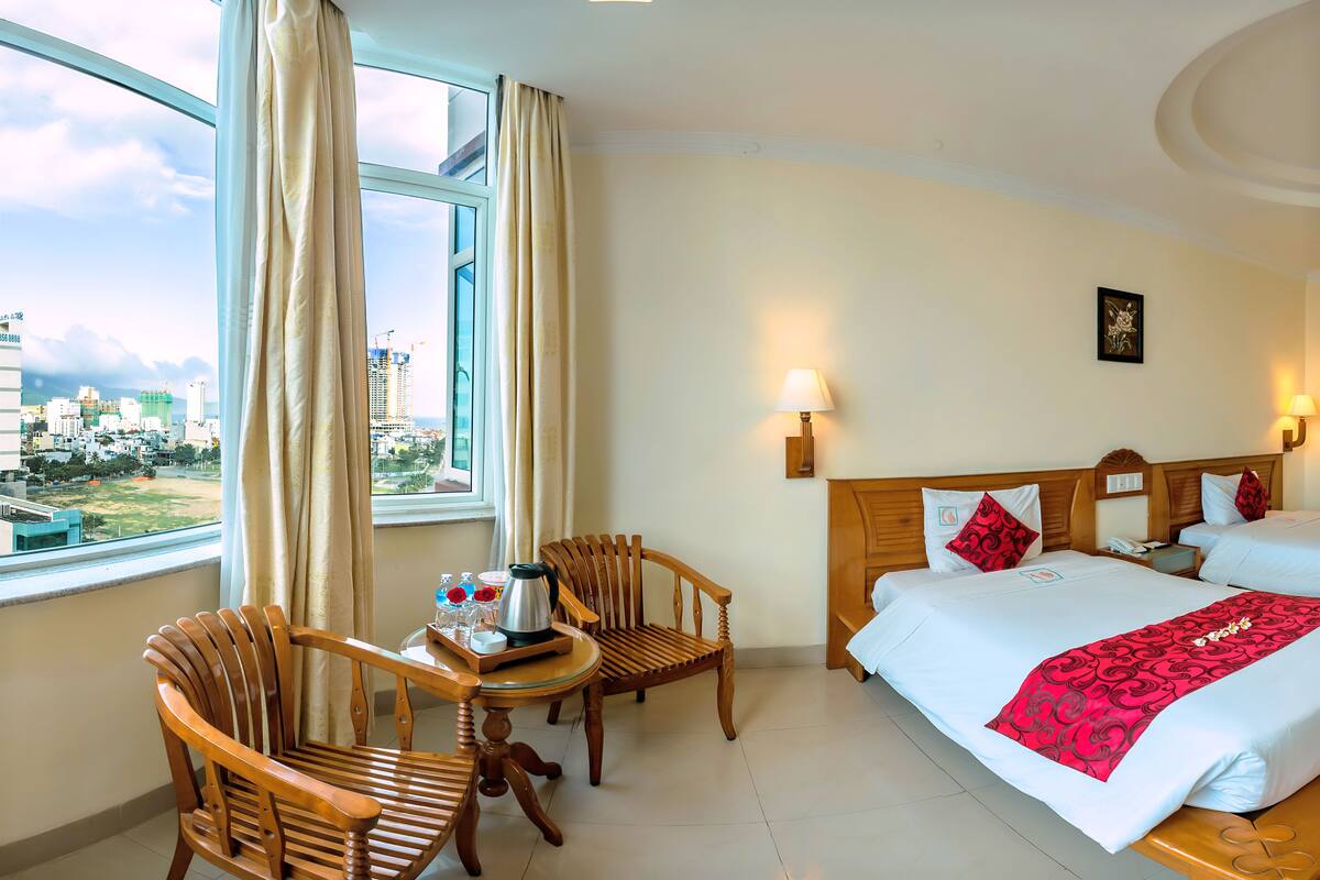 A hotel room with a large window, a bed with white linens and red accents, a wooden chair, a small table with a coffee pot, and a city view.