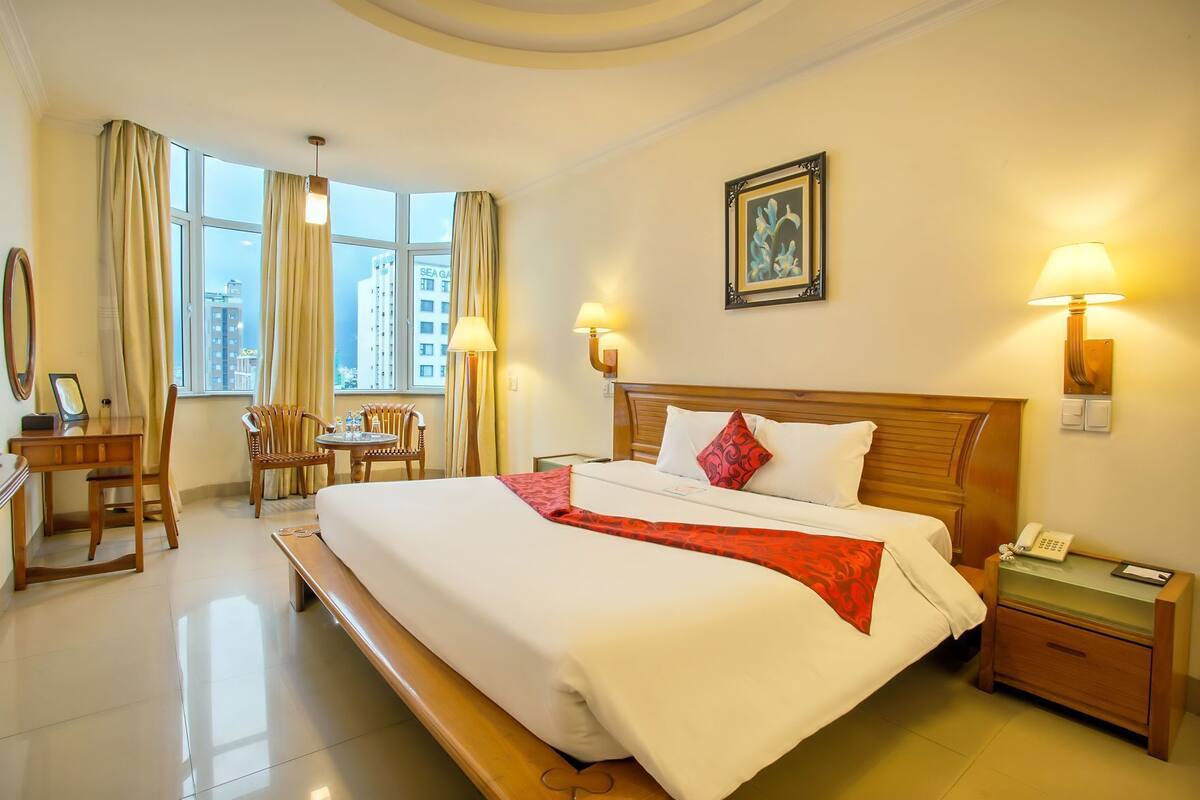 A hotel room with a large bed, bedside table, lamp, and a view of the cityscape through the window.