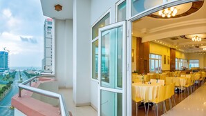 A hotel room with a balcony overlooking a cityscape, a dining area with yellow tables, and a view of a tall building labeled NHU. - Golden Sea Hotel (Da Nang)
