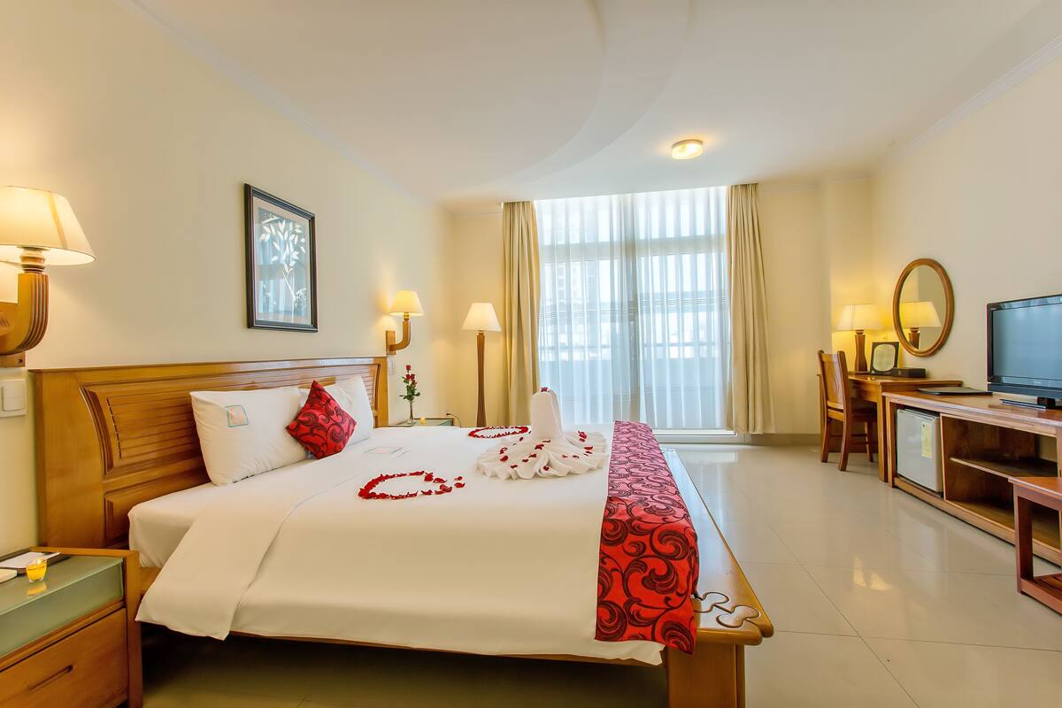 A hotel room with a bed, bedside lamps, a television, a desk, and a window with curtains.