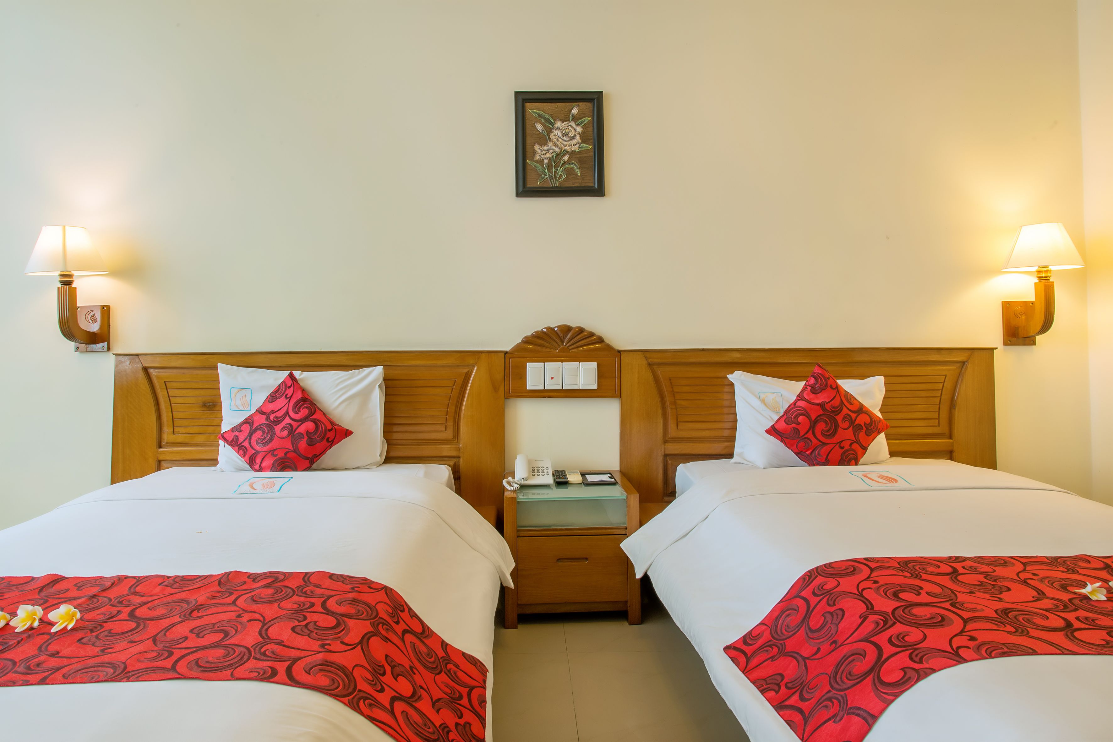 A hotel room with two beds, each with red and white bedding, a wooden headboard, bedside lamps, a nightstand with a phone, and a framed picture on the wall.