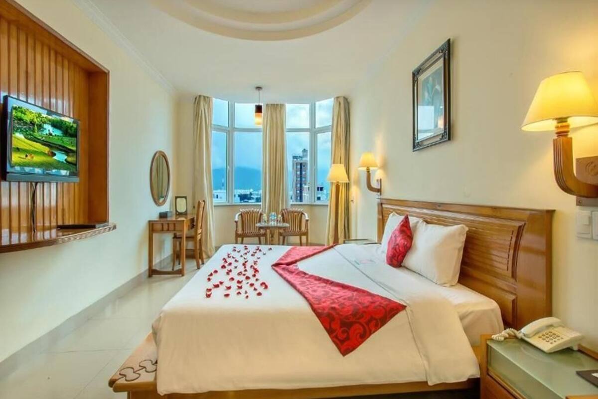 A hotel room with a large bed, a desk with a telephone, a TV mounted on the wall, and a view of the city through large windows.