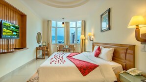 A hotel room with a large bed, a TV mounted on the wall, a desk with a lamp, a chair, and a view of the city through large windows. - Golden Sea Hotel (Da Nang)
