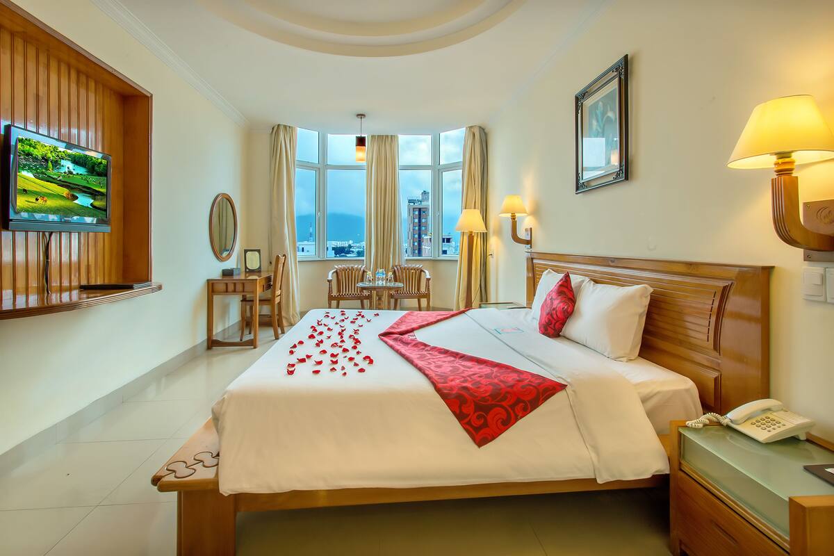 A hotel room with a large bed, a TV mounted on the wall, a desk with a lamp, a chair, and a view of the city through large windows.