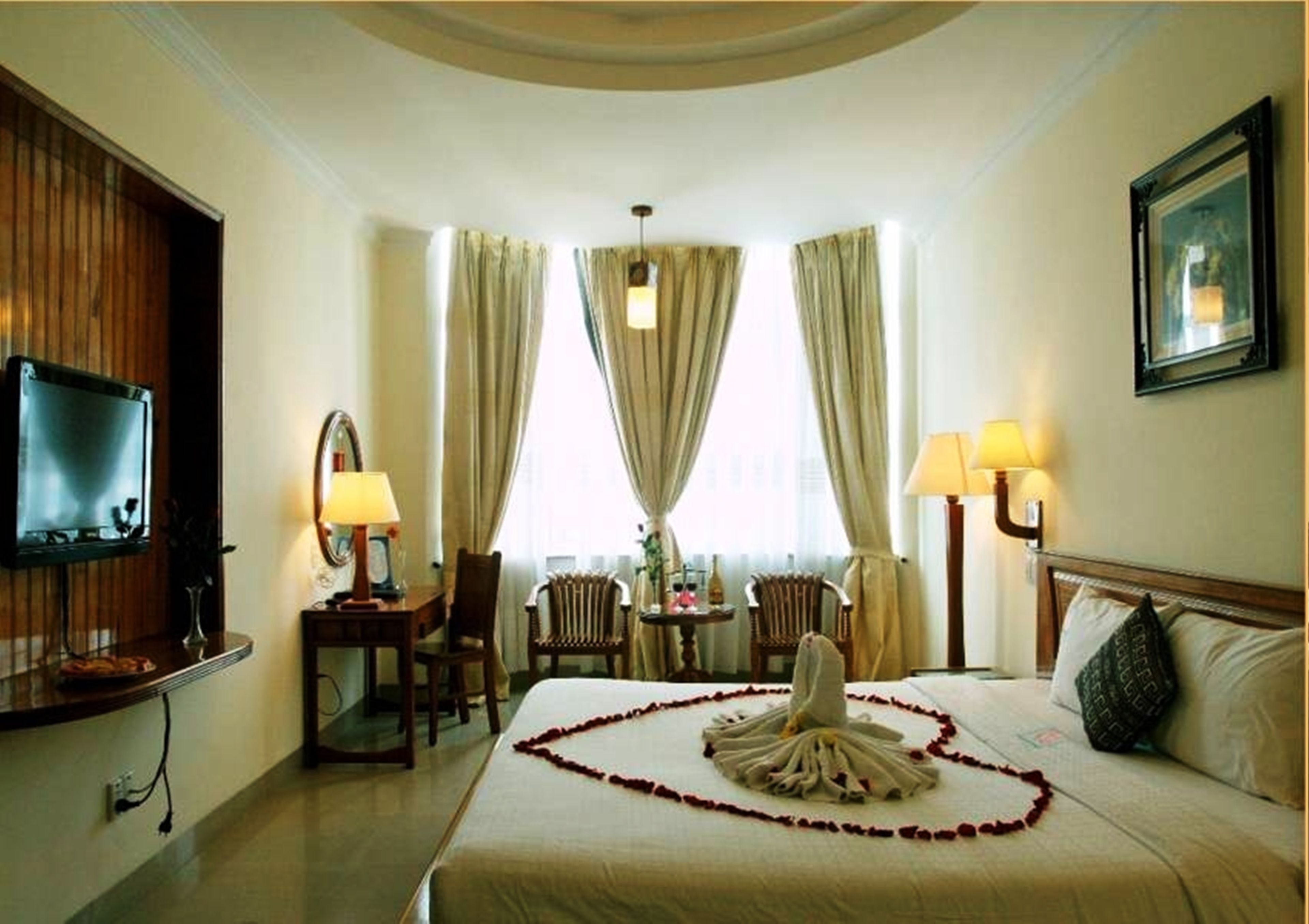 A hotel room with a bed, a TV, two lamps, a chair, a table, and a painting on the wall.