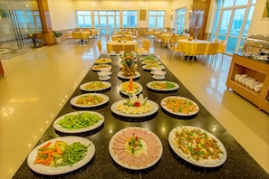 A buffet setup with various dishes arranged on a long table in a spacious room with multiple tables and chairs.