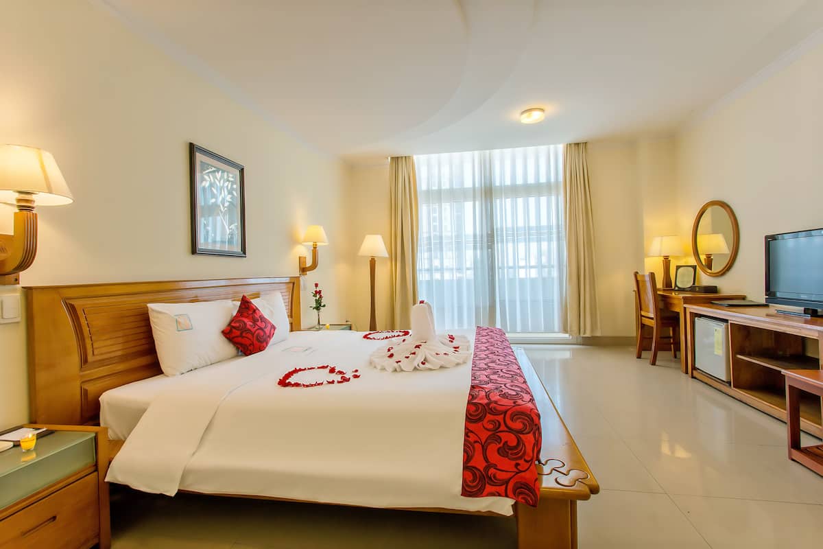 A hotel room with a neatly made bed, a desk with a computer, a television, and a large window with curtains.