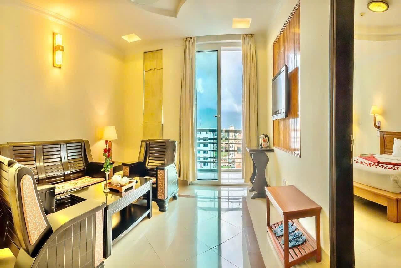 A hotel room with a sofa, a dining table, a chair, a small table, a bed, a TV, and a balcony with a view.