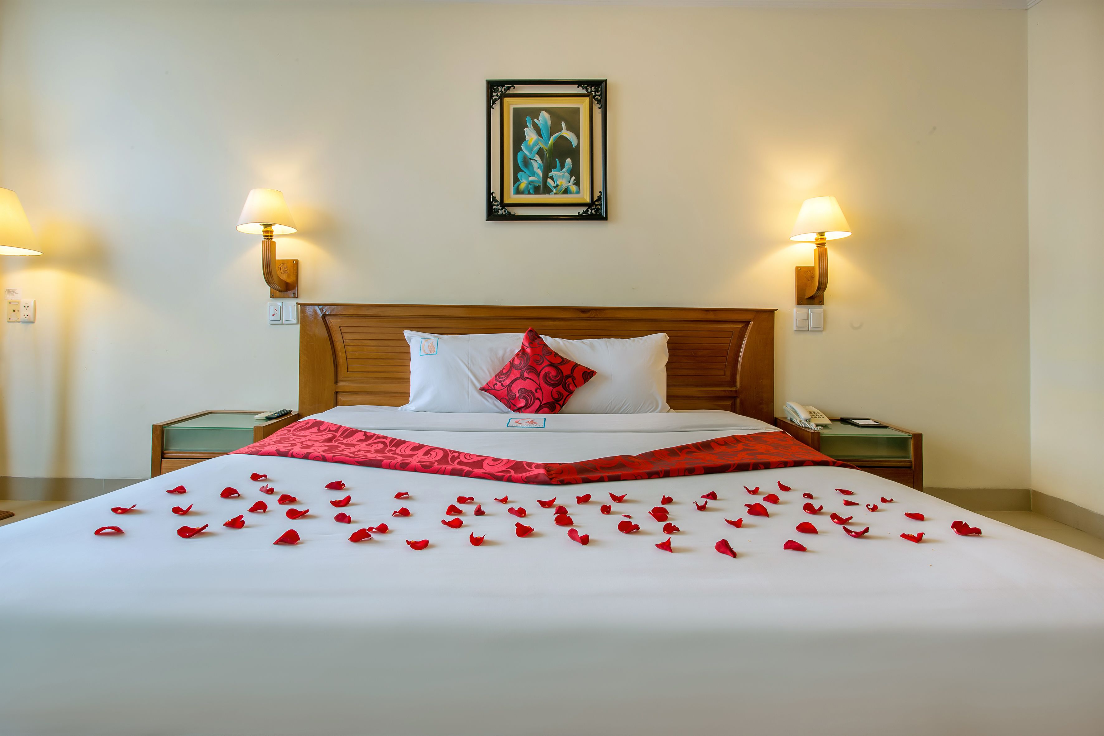 Una cama impecable con pétalos de rosa roja, un almohadón blanco y uno rojo. Hay dos mesitas de noche con lámparas y un cuadro en la pared.