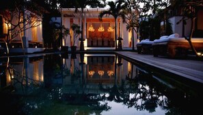 Un espace piscine paisible où se reflète l’intérieur du bâtiment, avec une terrasse en bois et des chaises longues.