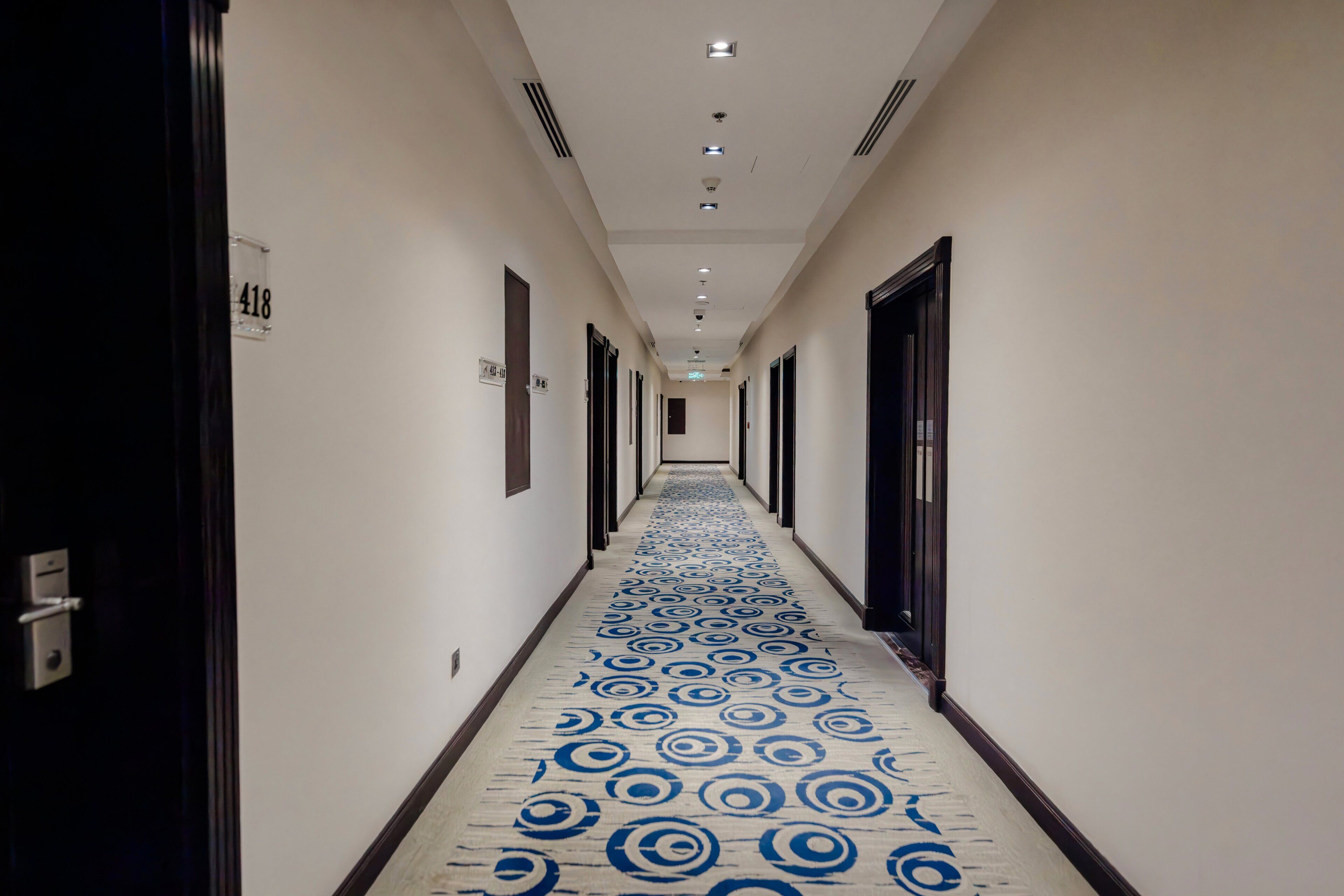 A hotel hallway with a patterned carpet, doors on both sides, and a number sign on the wall.
