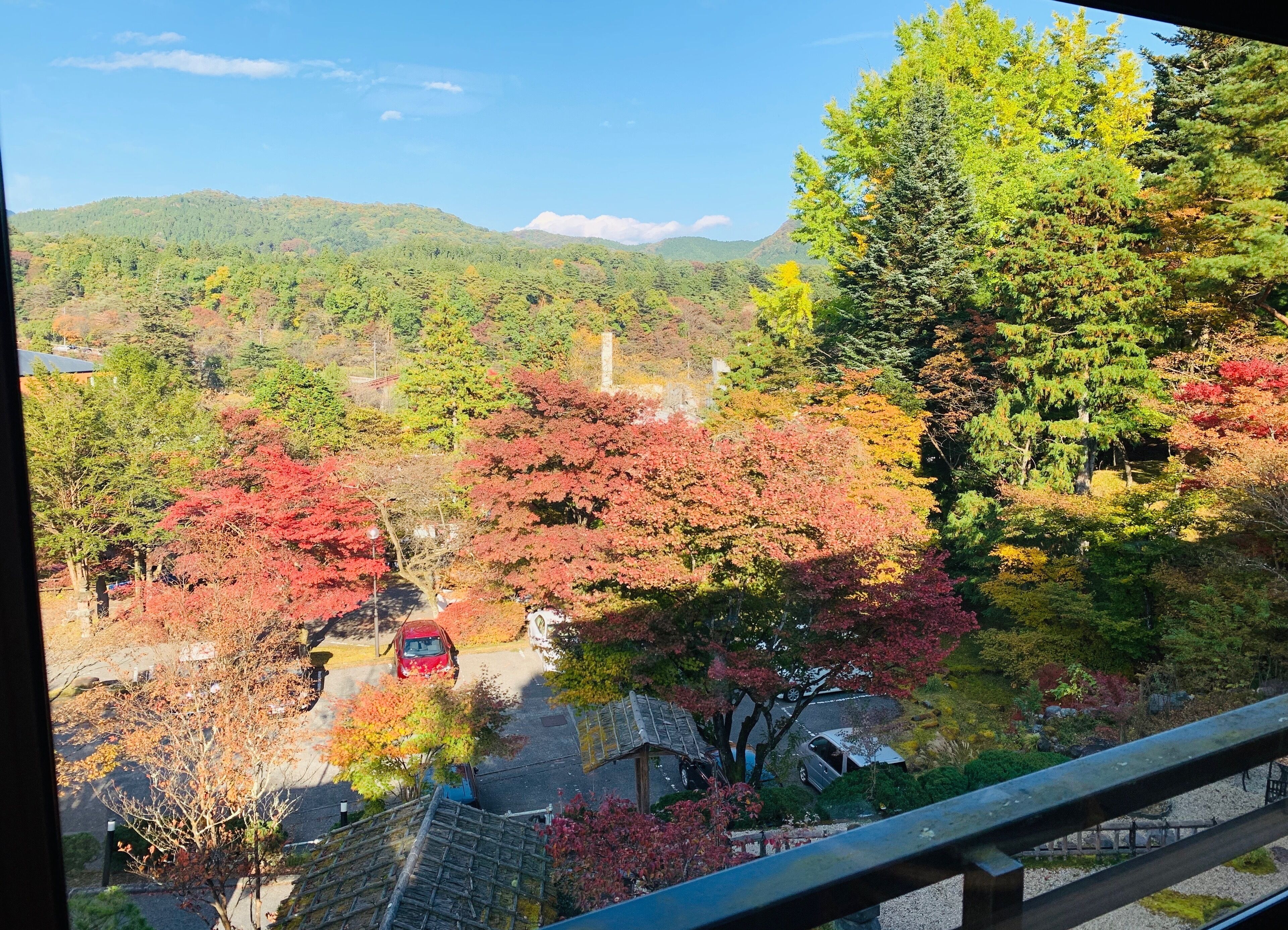 Vista de un aparcamiento con árboles mostrando colores otoñales, un vehículo rojo y un edificio tradicional japonés.