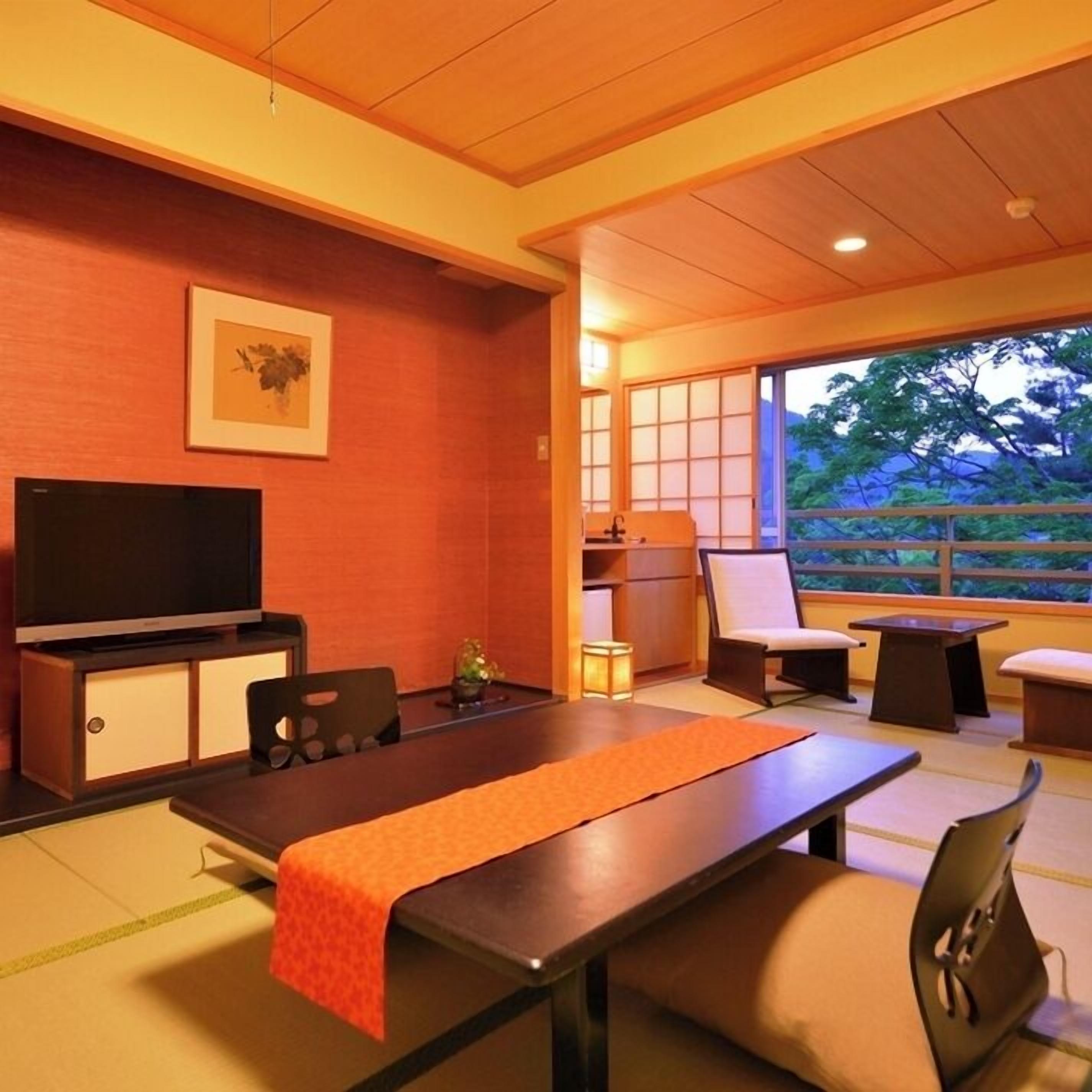 A room with a TV, a table with a red runner, chairs, a cabinet, and a view of greenery outside.