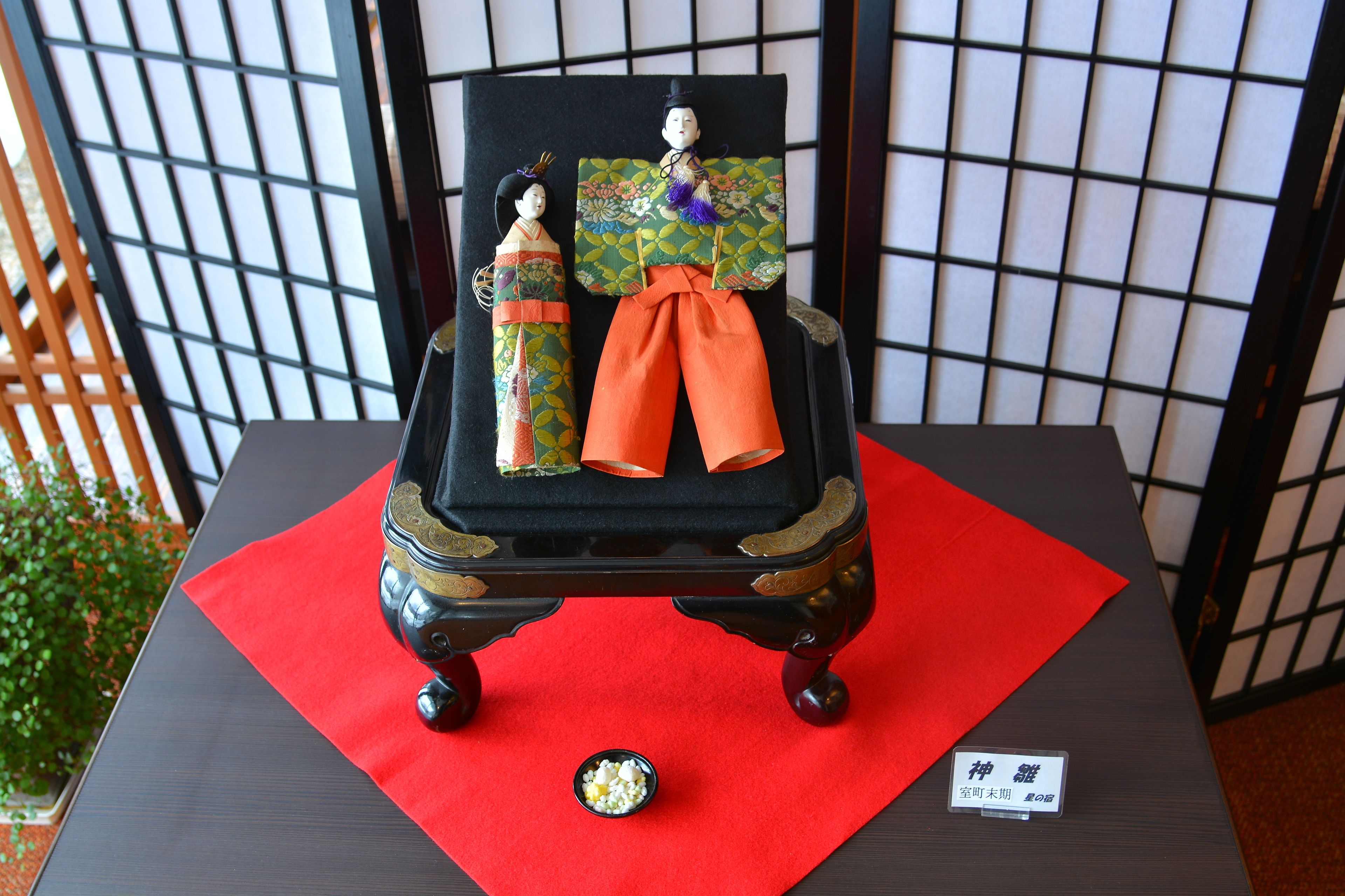 A traditional Japanese doll display on a table with a red cloth, set against a backdrop of a grid-patterned screen.