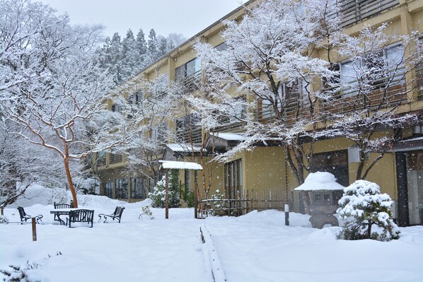 Un patio cubierto de nieve con bancos, un pequeño árbol y un edificio con varias ventanas.