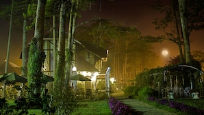 A nighttime view of a lodge with a patio area, surrounded by tall trees and a garden with purple flowers. - Cadasa Resort Dalat (Xuan Huong - Da Lat)
