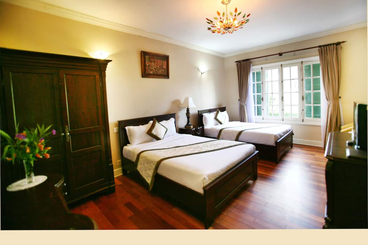 Habitación de hotel con dos camas, piso de madera, un candelabro, una ventana con cortinas y un cuadro en la pared.