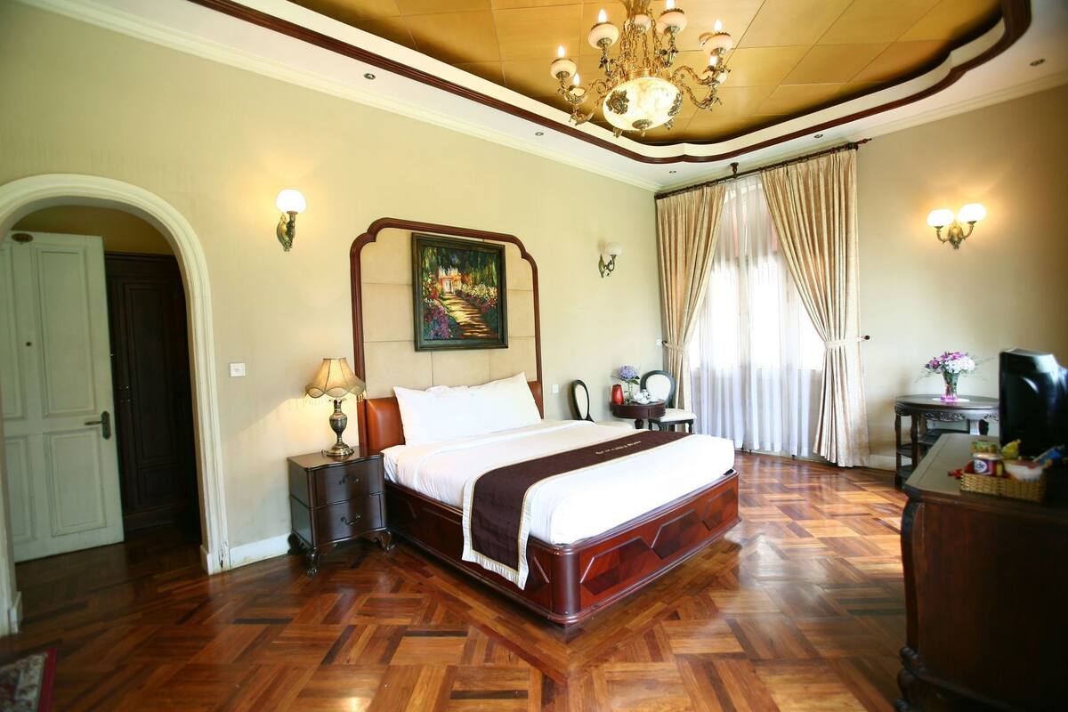 Habitación de hotel con una cama grande, suelo de madera, un candelabro, un cuadro, un escritorio con una computadora y una silla.