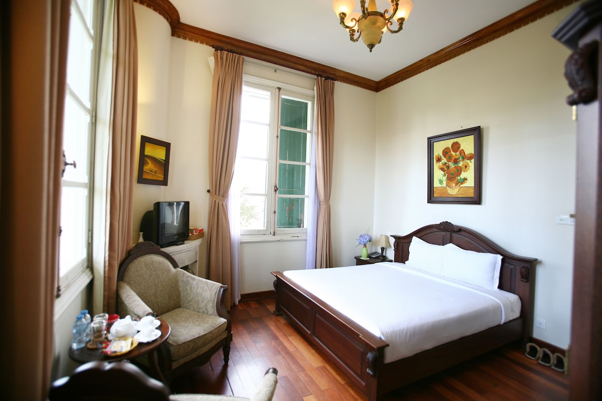 A hotel room with a bed, a chair, a small table with items, a television, a window with curtains, and a painting on the wall.