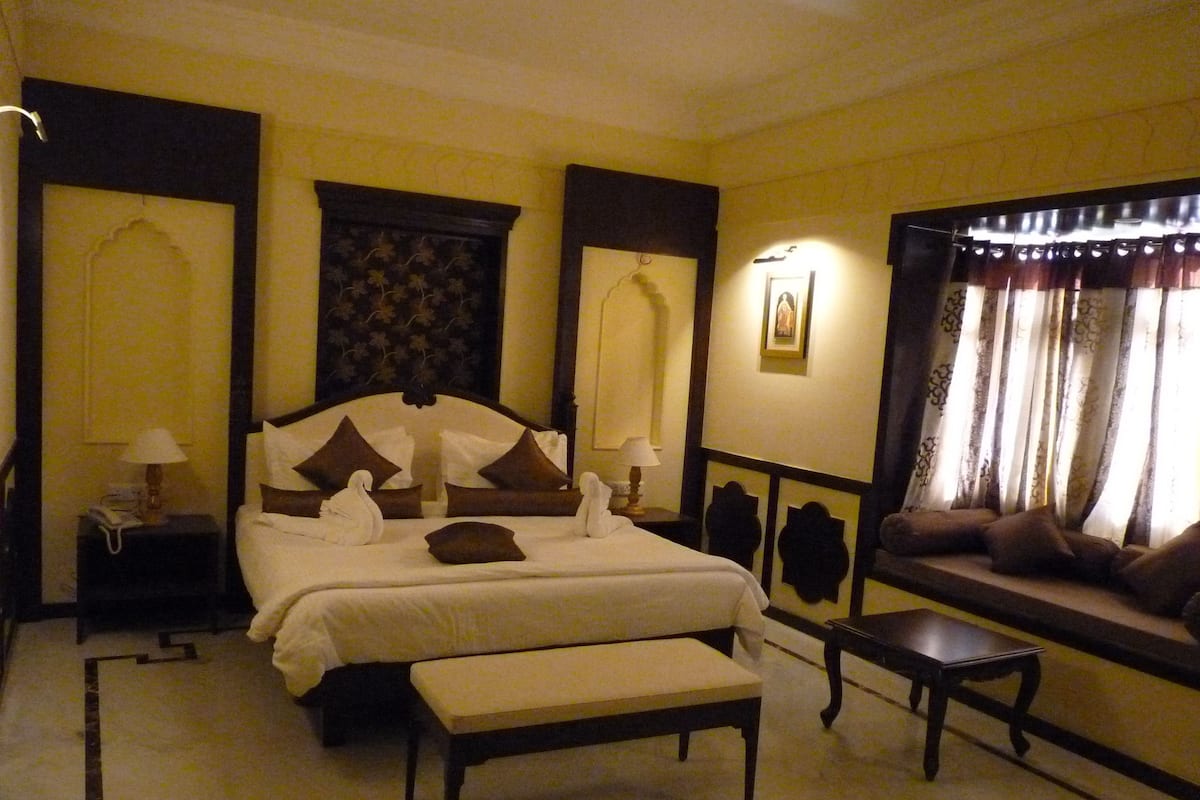 A hotel room with a large bed, two bedside tables, a bench, a sofa, and a window with curtains.