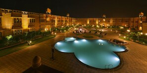Una piscina amplia e iluminada en el centro de un patio con un edificio imponente al fondo.