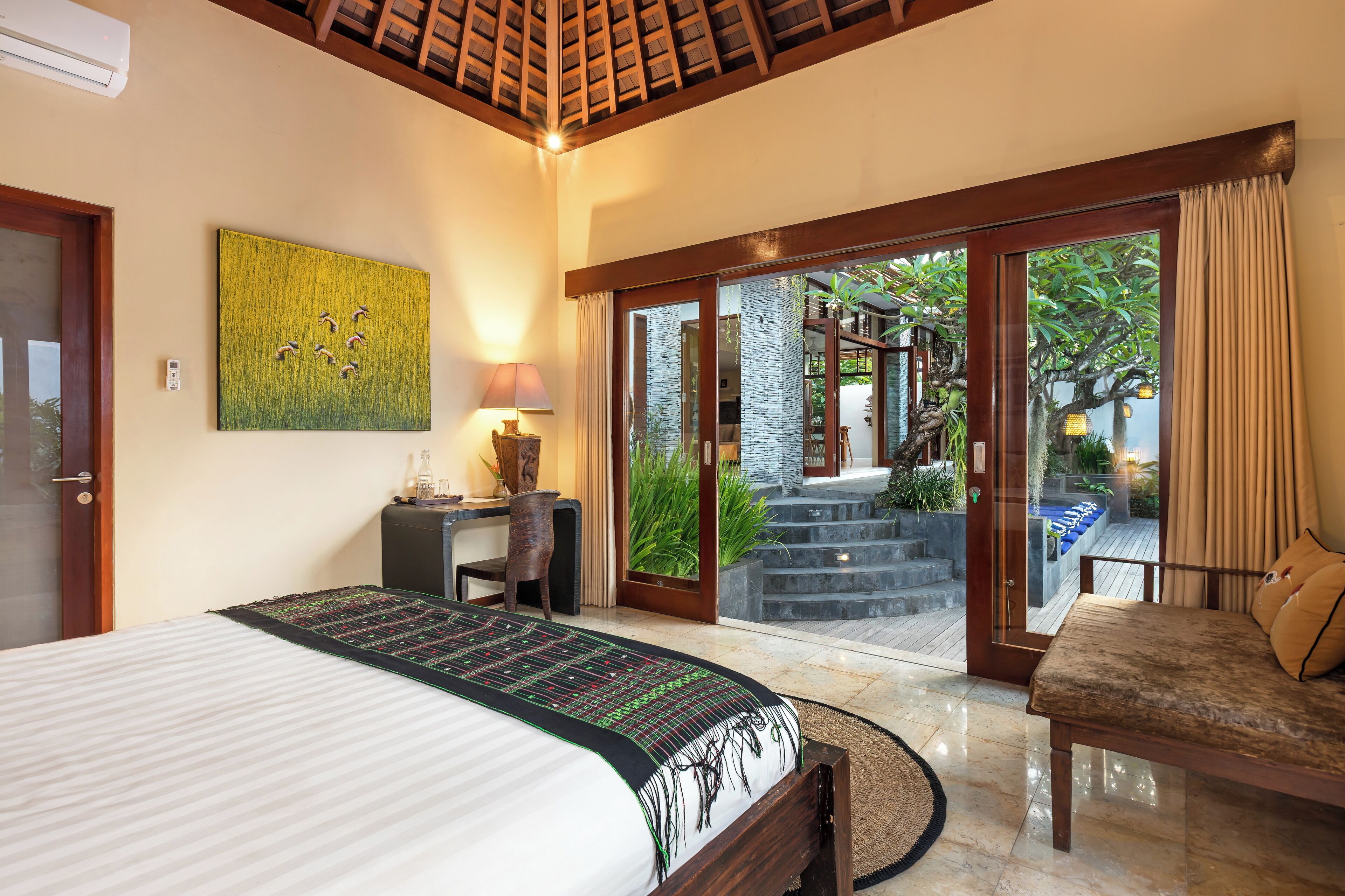 A bedroom with a bed, a desk, a chair, and a sofa. There is a sliding glass door leading to an outdoor area with plants and a staircase.
