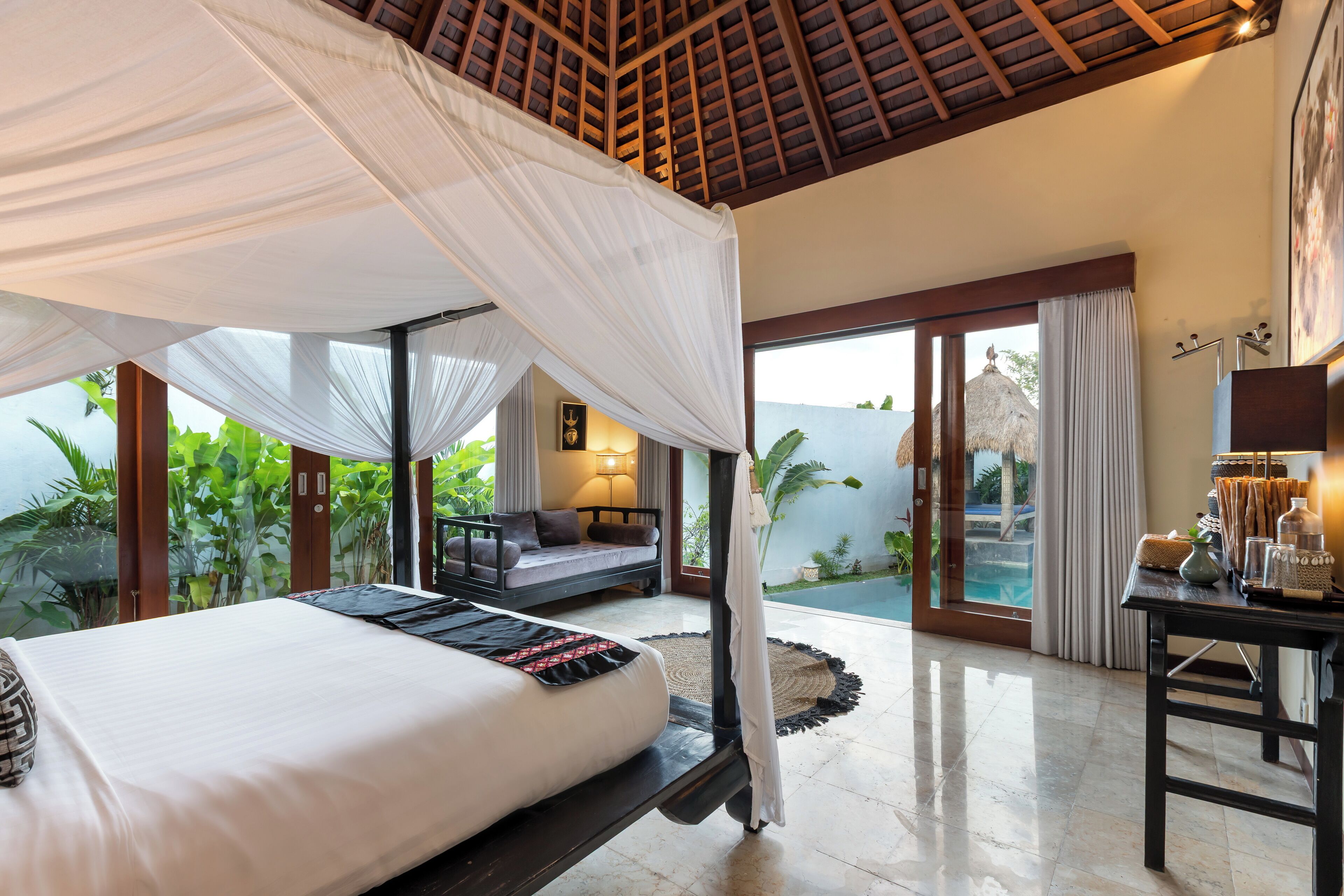 Un dormitorio con cama con dosel, una zona de estar con sofá, una mesa de comedor y vistas a la piscina y la vegetación.