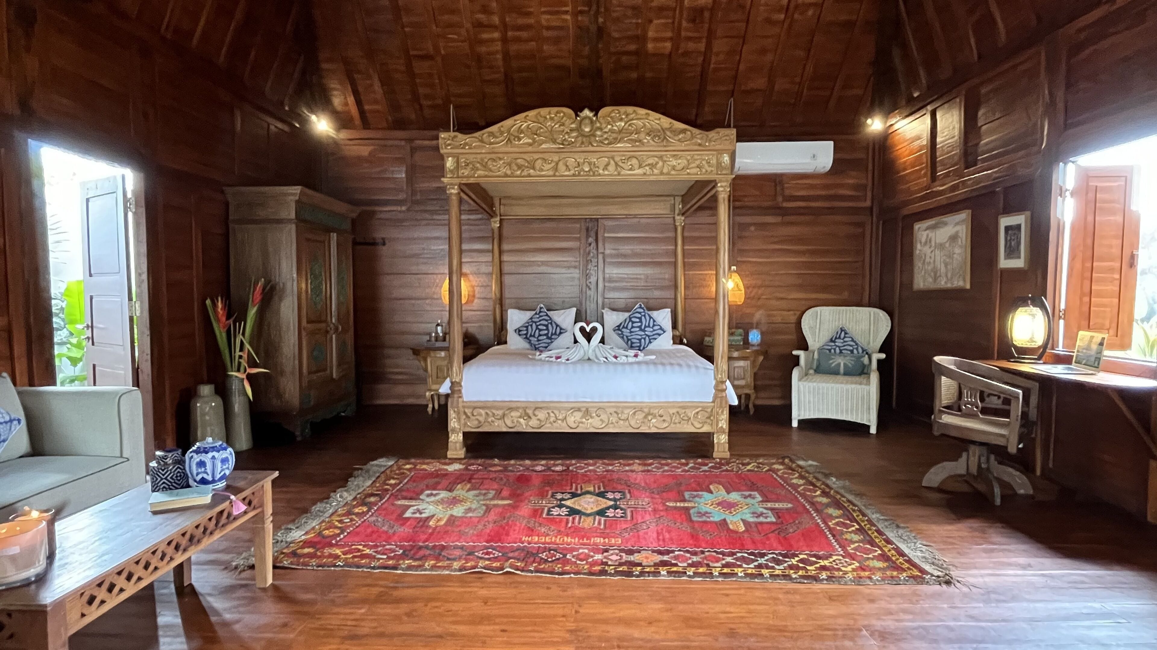 A four-poster bed with a red and gold patterned rug, wooden walls, and a small table with a lamp.