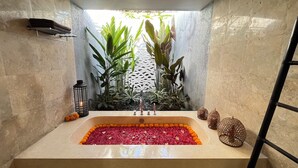Un baño con una bañera rectangular llena de pétalos de rosa, rodeado de plantas en macetas y un farol decorativo.