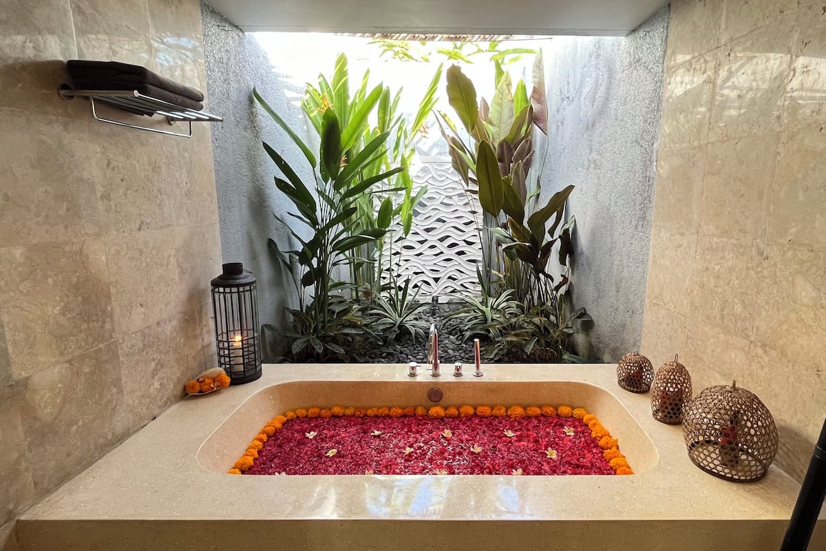 Ein Badezimmer mit einer rechteckigen Badewanne, gefüllt mit Rosenblüten, umgeben von Topfpflanzen und einem dekorativen Laternenleuchter.