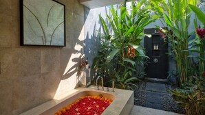 Un baño con una bañera grande llena de flores, una obra de arte en la pared y vistas a un jardín con plantas altas y una puerta.