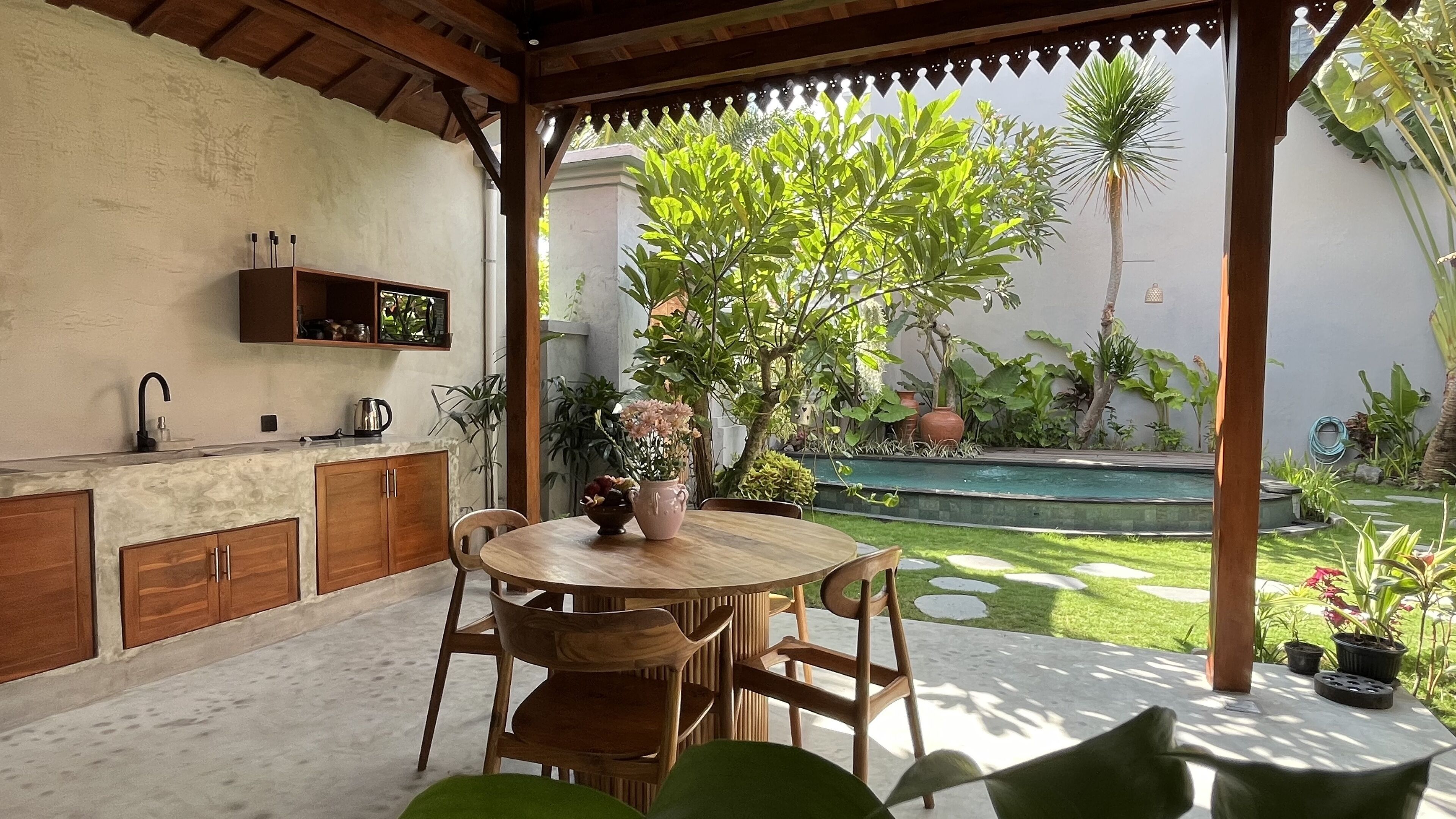 Une terrasse avec une table ronde en bois et des chaises, un petit coin cuisine avec un évier et des placards, et une piscine en arrière-plan.